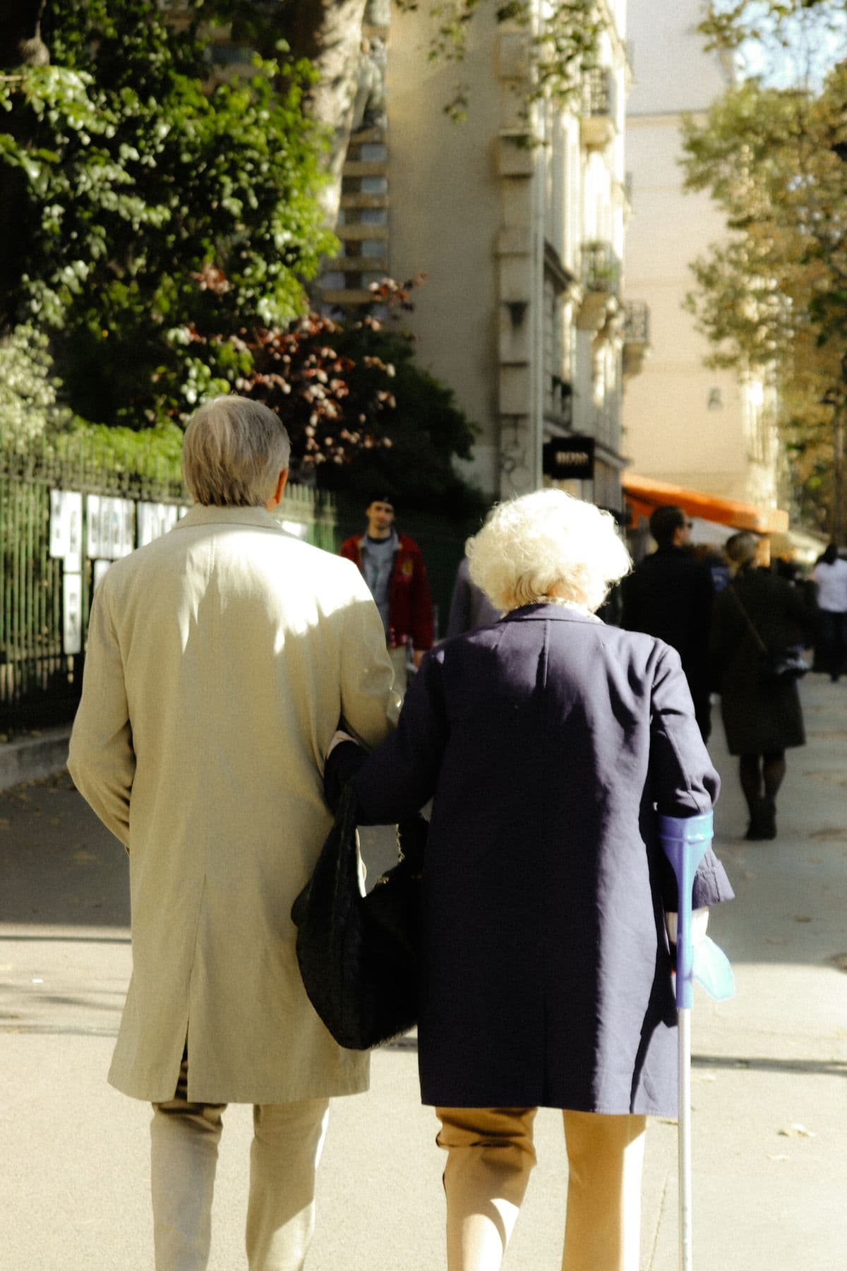 An older couple walking arm in arm along a sunny street, seen from behind — illustrative demo image